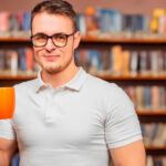 Homem com as mãos deformadas, em uma biblioteca, segurando uma xícara de café laranja